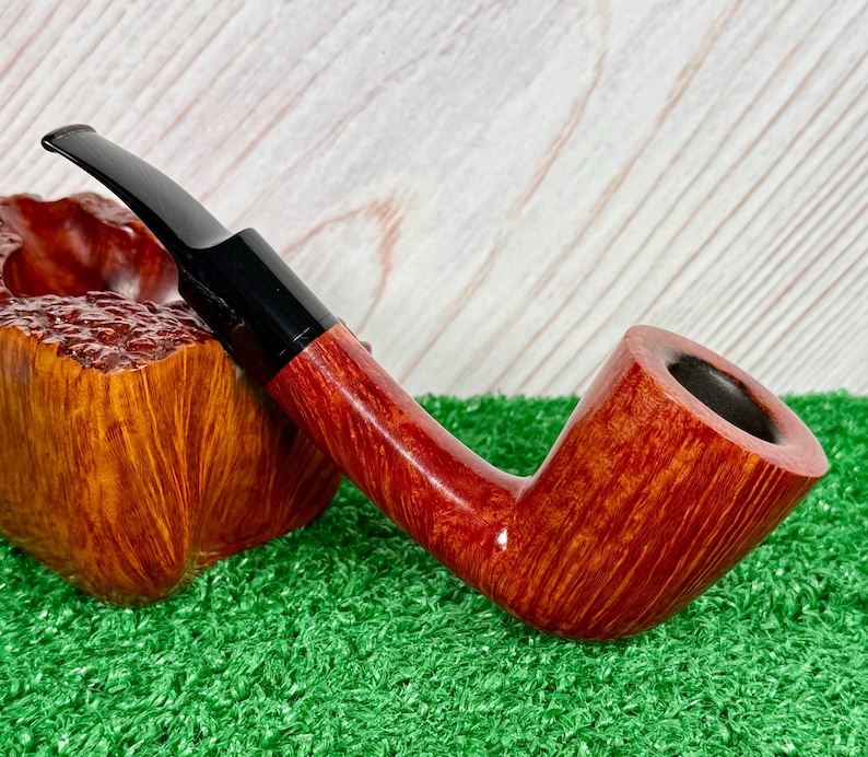 May include: A tobacco pipe with a polished reddish-brown wooden bowl and a smooth black acrylic stem. The pipe features a distinct grain pattern on the wood and is positioned next to a textured, burl-like wooden block with a deep amber finish.