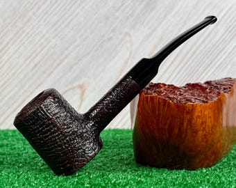 May include: A tobacco pipe with a dark brown, textured bowl and a long, smooth black stem. The pipe is positioned next to a piece of raw, polished briar wood that displays a natural, light brown grain pattern and a glossy, uneven surface.