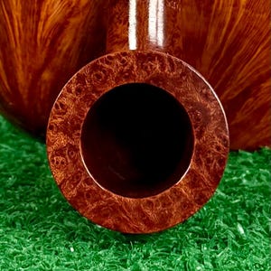 May include: A polished brown tobacco pipe with a prominent circular bowl opening and a smooth, swirling wood grain pattern. The stem features the engraved text KAYWOODIE, showcasing a rich, glossy finish and a deep, hollow chamber.