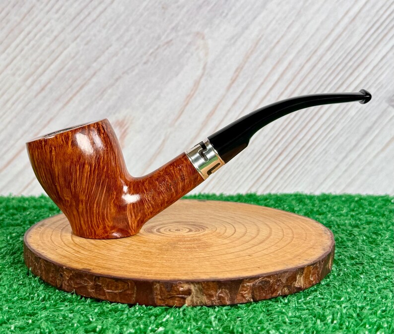 May include: A tobacco pipe with a polished light brown wooden bowl featuring a visible grain pattern. The pipe has a curved black stem connected to the bowl by a silver-colored metal band with a geometric cutout design detail.