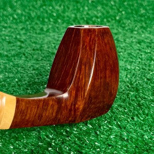 Pode incluir: Um cachimbo de tabaco de madeira castanha polida com um acabamento de gr&atilde;o escuro e liso e uma forma de fornilho facetada distinta. O cachimbo apresenta uma sec&ccedil;&atilde;o de haste de madeira de cor mais clara perto da boquilha, com textura natural.