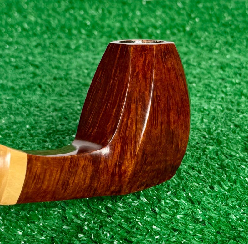 Pode incluir: Um cachimbo de tabaco de madeira castanha polida com um acabamento de gr&atilde;o escuro e liso e uma forma de fornilho facetada distinta. O cachimbo apresenta uma sec&ccedil;&atilde;o de haste de madeira de cor mais clara perto da boquilha, com textura natural.