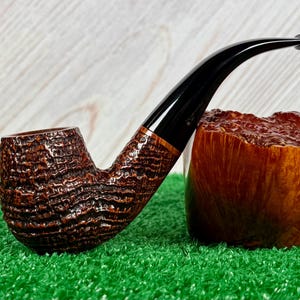 May include: A brown tobacco pipe with a textured bowl and a curved black stem. The pipe has a copper-colored band near the stem's base. The pipe rests on a green surface, with a wooden block in the background.