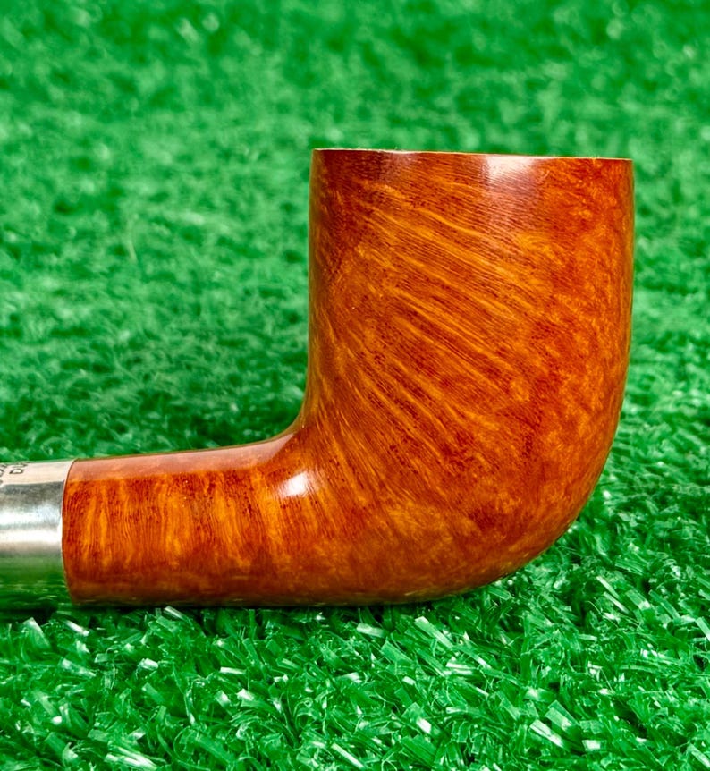 Puede incluir: Una pipa de tabaco con un hornillo de madera pulida de color marr&oacute;n claro con un patr&oacute;n de veta natural visible. La pipa incluye una banda de color plata met&aacute;lica que conecta el hornillo con la boquilla, con un dise&ntilde;o liso y curvo.