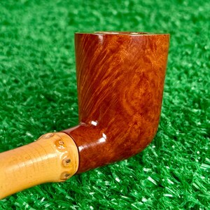 May include: A handcrafted tobacco pipe with a curved, polished, brown bowl and a light-colored, bamboo-style stem. The bowl has a rich, wood-grain pattern. The pipe rests on a green surface.