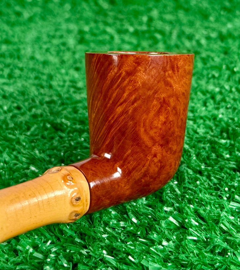May include: A handcrafted tobacco pipe with a curved, polished, brown bowl and a light-colored, bamboo-style stem. The bowl has a rich, wood-grain pattern. The pipe rests on a green surface.