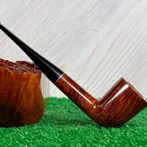 May include: A tobacco pipe with a polished brown wooden bowl featuring a visible grain pattern and a long, slender black stem. The stem is connected to the bowl by a thin silver-colored metal band. The pipe is positioned at an angle.