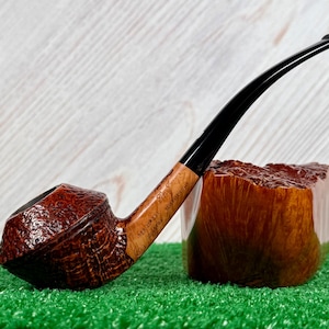 Pode incluir: Um cachimbo de tabaco castanho com uma haste preta curva repousa sobre uma superfície verde. O cachimbo tem uma tigela texturizada e uma haste de madeira clara com detalhes gravados. Um suporte de cachimbo de madeira está ao lado do cachimbo.