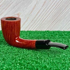 May include: A tobacco pipe featuring a polished, reddish-brown wooden bowl with a visible grain pattern and a smooth, black acrylic stem. The pipe has a curved shape and a cylindrical chamber opening at the top, showcasing a classic design.