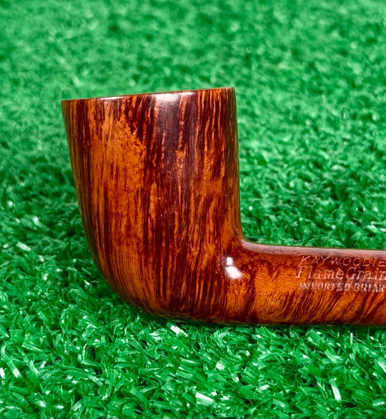 May include: A brown tobacco pipe made of imported briar wood with a distinct flame grain pattern. The side of the pipe is stamped with the text Kaywoodie Flame Grain IMPORTED BRIAR in a clear, legible font against the polished wood surface.