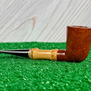 May include: A tobacco pipe with a dark brown bowl and a light brown bamboo-style stem. The mouthpiece is black. The pipe rests on a green surface with a light wood background.