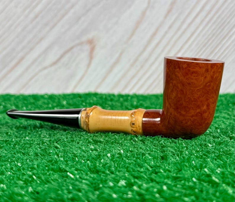 May include: A tobacco pipe with a dark brown bowl and a light brown bamboo-style stem. The mouthpiece is black. The pipe rests on a green surface with a light wood background.