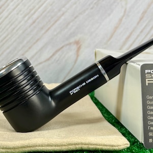 Pode incluir: Cachimbo Porsche Design P'3611 preto com um detalhe prateado. O cachimbo tem uma piteira canelada e um tubo longo e fino. A caixa ao lado tem o texto "PORSCHE SMOKING P'3611".