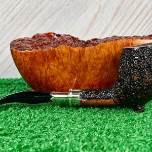 May include: A tobacco pipe with a dark, textured bowl and a black stem. The pipe rests on a bed of green artificial grass, with a wooden bowl in the background. The pipe has a silver band.