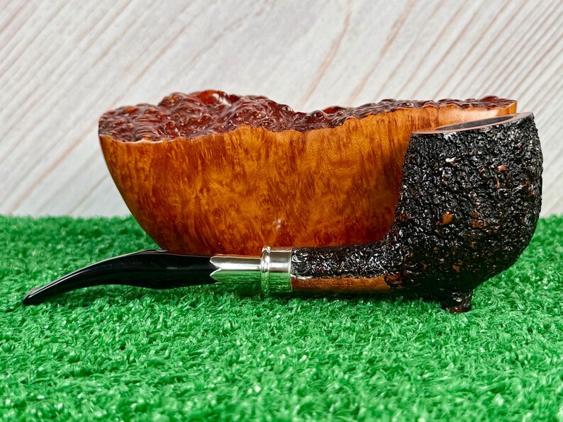 May include: A tobacco pipe with a dark, textured bowl and a black stem. The pipe rests on a bed of green artificial grass, with a wooden bowl in the background. The pipe has a silver band.