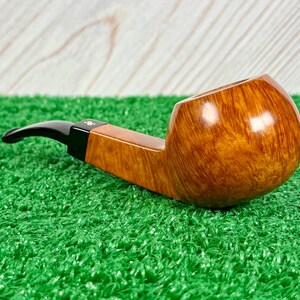 May include: A tobacco pipe with a polished, light brown, natural wood grain bowl and a smooth, black acrylic stem. The stem features a small, white, stylized logo symbol near the junction where it connects to the wooden shank of the pipe.