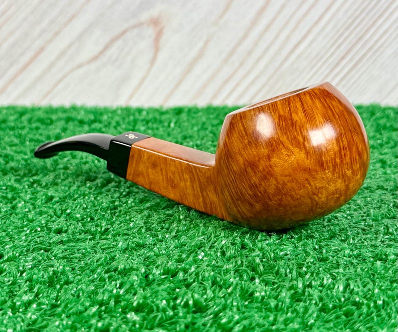 May include: A tobacco pipe with a polished, light brown, natural wood grain bowl and a smooth, black acrylic stem. The stem features a small, white, stylized logo symbol near the junction where it connects to the wooden shank of the pipe.