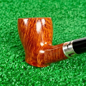 May include: A tobacco pipe with a polished, light brown wood grain bowl featuring a visible engraved signature on the side. The pipe includes a silver-colored metal band connecting the bowl to a smooth, black stem, showcasing a classic design.