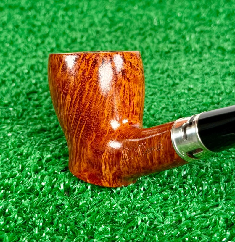 May include: A tobacco pipe with a polished, light brown wood grain bowl featuring a visible engraved signature on the side. The pipe includes a silver-colored metal band connecting the bowl to a smooth, black stem, showcasing a classic design.