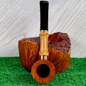 May include: A wooden tobacco pipe with a black stem and a light brown bowl. The pipe has a decorative band near the stem and is set against a wooden background. The pipe is resting on a green surface.