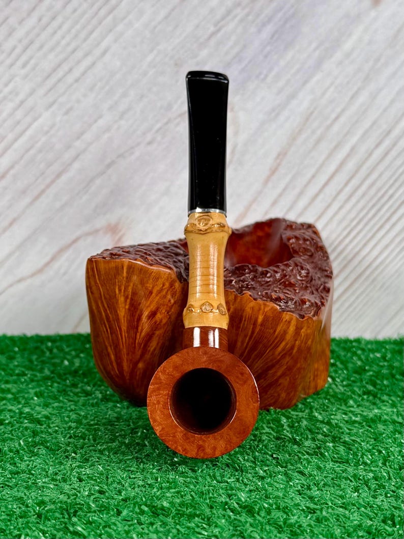 May include: A wooden tobacco pipe with a black stem and a light brown bowl. The pipe has a decorative band near the stem and is set against a wooden background. The pipe is resting on a green surface.