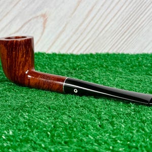 May include: A brown wooden tobacco pipe with a smooth finish and a long, black acrylic stem. The bowl features a visible wood grain pattern. The stem is marked with a small white circular inlay and the text Kaywoodie Fine Grain Imported.