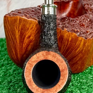 May include: Close-up of a tobacco pipe with a black, textured bowl and a silver band. The pipe's rim is a light brown color, and the interior is black. The background features a wooden pipe stand and green artificial turf.