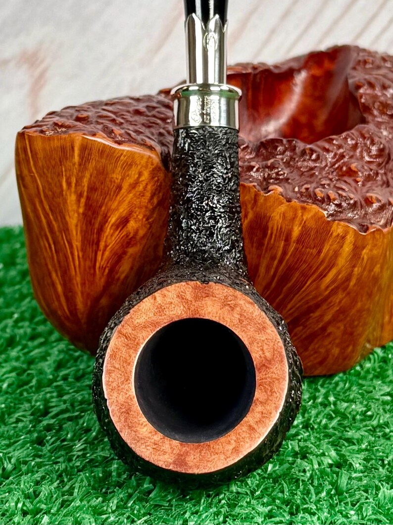 May include: Close-up of a tobacco pipe with a black, textured bowl and a silver band. The pipe's rim is a light brown color, and the interior is black. The background features a wooden pipe stand and green artificial turf.