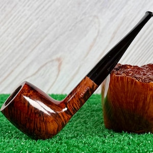 Puede incluir: Una pipa de tabaco clásica con un cuenco de madera curvado y pulido y una boquilla larga y negra. El cuenco tiene un rico patrón de vetas marrón oscuro. La pipa descansa sobre una superficie verde, con un recipiente de madera en el fondo.