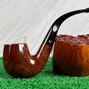 Pode incluir: Um cachimbo de tabaco curvo de madeira, com uma haste escura e brilhante e uma piteira arredondada. O cachimbo é castanho escuro com grão de madeira visível. A piteira é de um tom castanho mais claro e tem uma superfície texturizada. Uma pequena lua crescente branca é visível na haste.