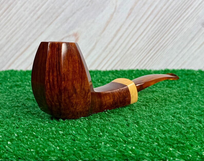 Pode incluir: Um cachimbo de tabaco com fornilho de madeira castanho-escuro polido e design facetado. A piteira &eacute; feita de um material transl&uacute;cido de cor &acirc;mbar e est&aacute; ligada ao fornilho por um anel de madeira clara e lisa que acentua a transi&ccedil;&atilde;o.