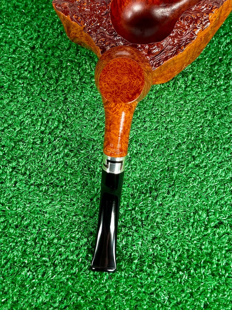 May include: A tobacco pipe featuring a polished brown briar wood bowl with a distinct burl grain pattern. The stem is crafted from a lighter-toned wood, connected to a black acrylic mouthpiece by a silver-colored metal band with a geometric cutout.