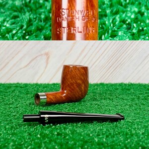 Op de afbeelding: Een bruine houten tabakspijp met een gladde afwerking en een zilveren band. De pijp is gegraveerd met de tekst STANWELL DANISH DESIGN STERLING. Het bevat een afneembare zwarte steel met een wit S-logo bij het mondstuk.