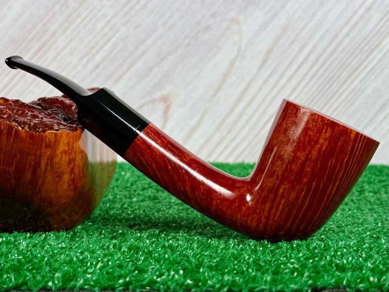 May include: A tobacco pipe with a polished reddish-brown wooden bowl and a smooth, curved black stem. The pipe features a visible wood grain pattern on the bowl and a sleek, tapered mouthpiece, showcasing a classic design for smoking tobacco.