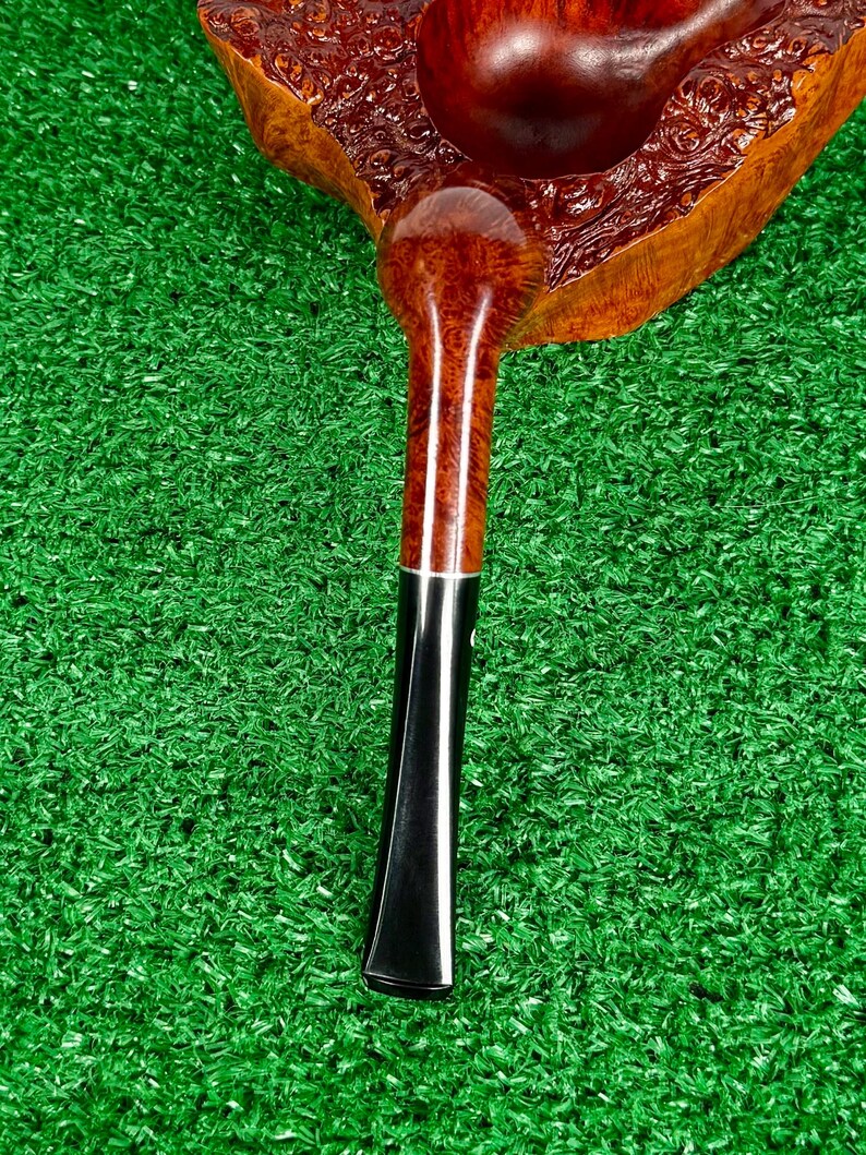 May include: A tobacco pipe featuring a polished, reddish-brown briar wood bowl with a distinct burl grain pattern. The stem is crafted from smooth, dark black material, connected to the bowl by a thin, silver-colored metal band at the junction.