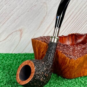 May include: A black and brown tobacco pipe with a textured bowl and a curved, glossy black stem. The pipe has a silver band and a small blue accent. The pipe rests on a green surface, with a wooden bowl in the background.