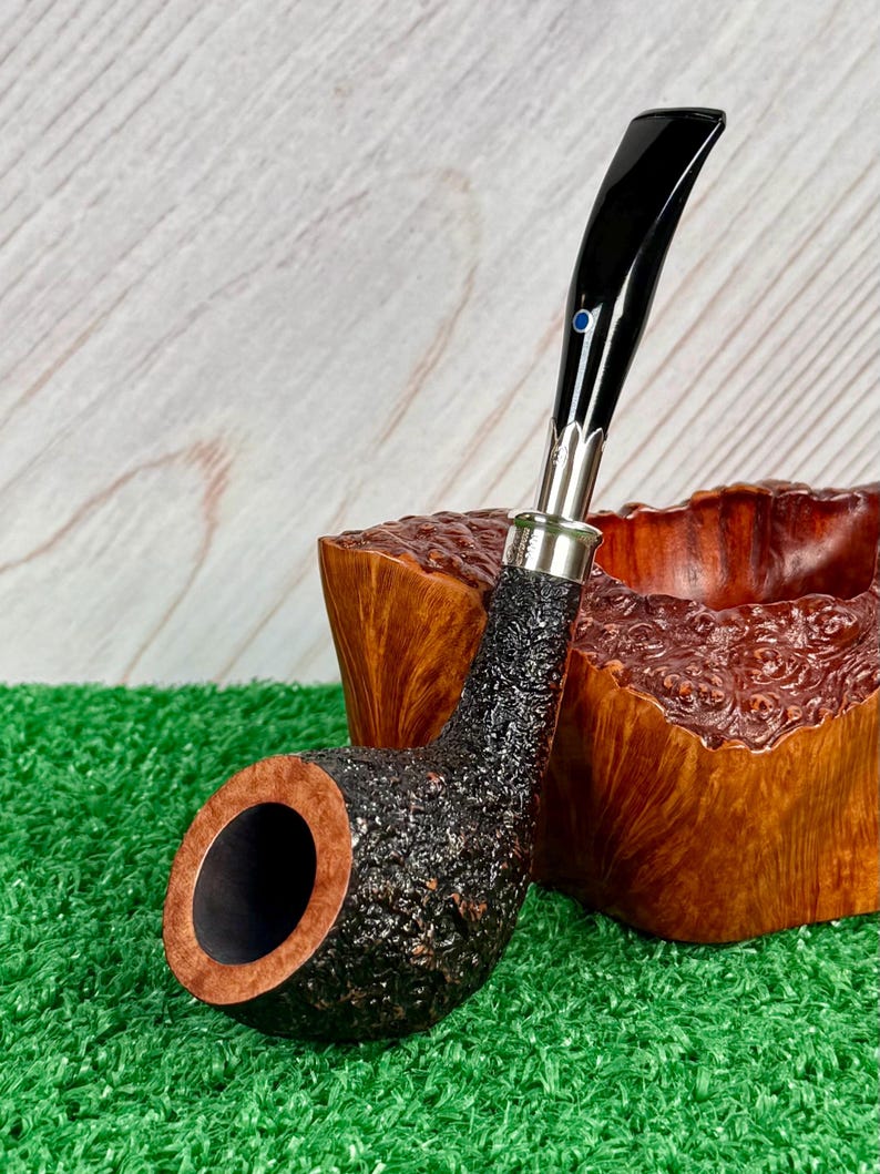 May include: A black and brown tobacco pipe with a textured bowl and a curved, glossy black stem. The pipe has a silver band and a small blue accent. The pipe rests on a green surface, with a wooden bowl in the background.