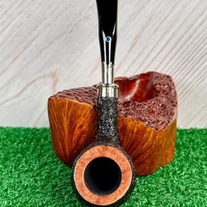 May include: A tobacco pipe with a black stem and a textured, dark brown bowl. The bowl has a light brown interior and is set on a wooden stand. A small blue accent is on the stem.