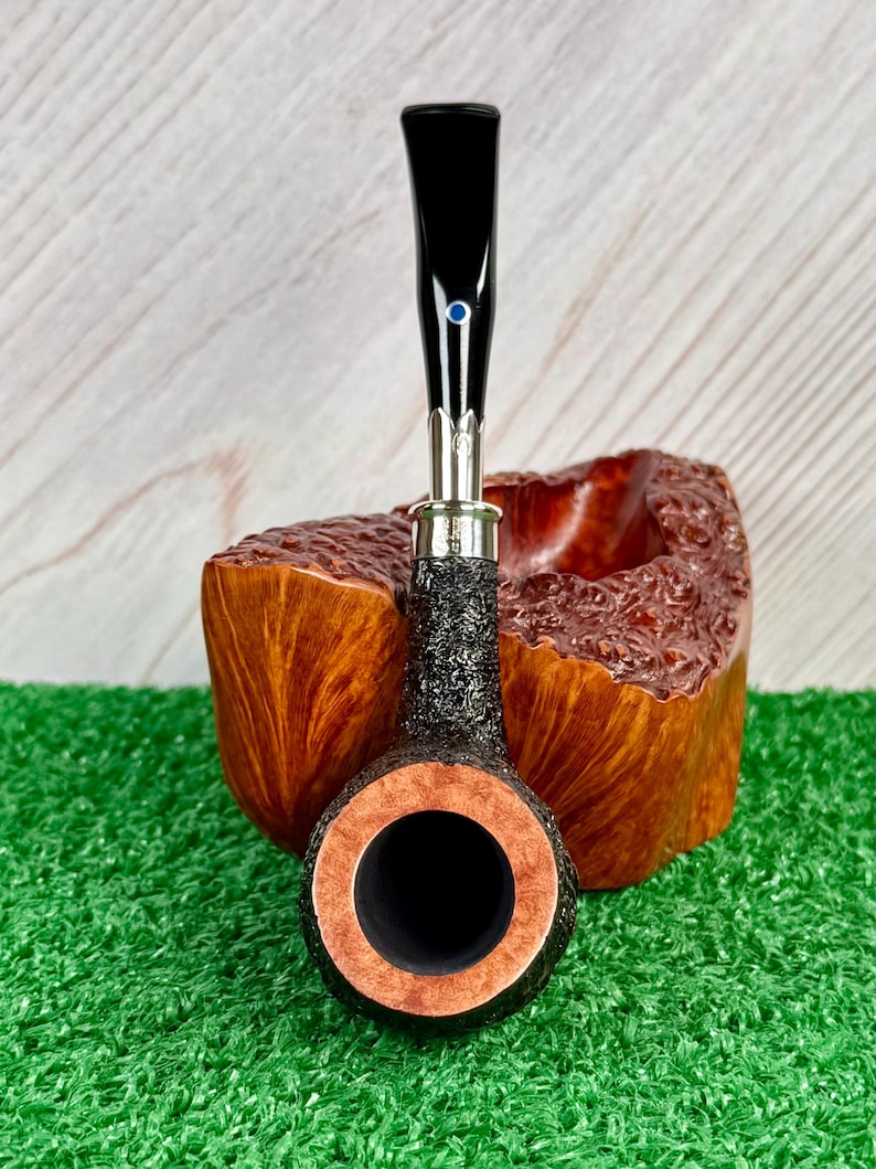 May include: A tobacco pipe with a black stem and a textured, dark brown bowl. The bowl has a light brown interior and is set on a wooden stand. A small blue accent is on the stem.