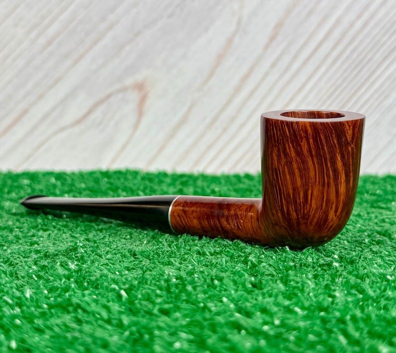May include: A tobacco pipe featuring a polished brown wooden bowl with a visible grain pattern and a long, smooth black stem. The pipe is constructed with a cylindrical bowl and a slender, tapered mouthpiece, showcasing a contrast between the natural wood finish and the dark, solid-colored stem material.