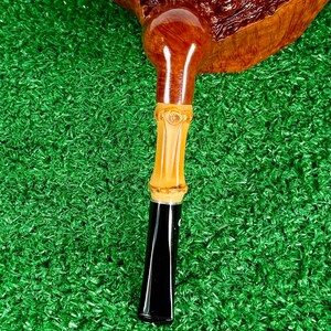 May include: A handcrafted tobacco pipe with a dark brown bowl and a light brown bamboo stem. The stem has a black mouthpiece. The pipe rests on a green surface.