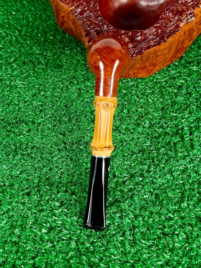May include: A handcrafted tobacco pipe with a dark brown bowl and a light brown bamboo stem. The stem has a black mouthpiece. The pipe rests on a green surface.