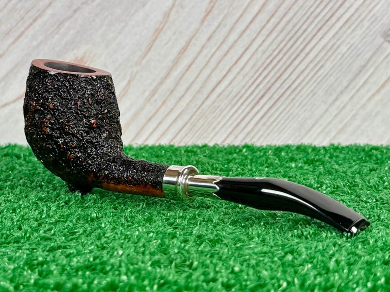 May include: A dark brown tobacco pipe with a textured bowl and a curved, glossy black stem. The pipe features a silver band and rests on a green surface, suggesting a classic smoking accessory.