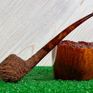 May include: A brown tobacco pipe with a long, slender stem and a textured bowl. The pipe rests on a green surface, with a wooden block beside it. The stem has a marbled appearance.