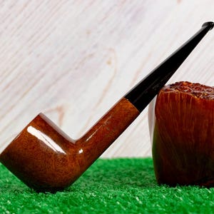 May include: A tobacco pipe with a polished brown wooden bowl and a long, slender black stem. The pipe is positioned next to a large, textured, reddish-brown wooden block, showcasing a smooth finish on the bowl and a dark mouthpiece.