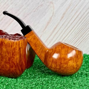 May include: A tobacco pipe with a polished light brown wooden bowl featuring a visible grain pattern and a smooth, rounded shape. The pipe has a slender, angled stem made of black material that extends from the bowl, showcasing a contrast in texture.