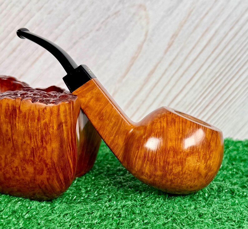 May include: A tobacco pipe with a polished light brown wooden bowl featuring a visible grain pattern and a smooth, rounded shape. The pipe has a slender, angled stem made of black material that extends from the bowl, showcasing a contrast in texture.