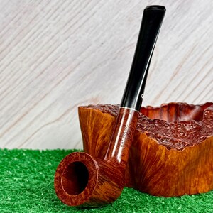 May include: A brown wooden tobacco pipe with a smooth finish and a black stem, featuring the text Kaywoodie stamped on the side. The pipe is positioned in front of a textured, natural-edged wooden bowl with a deep reddish-brown grain.