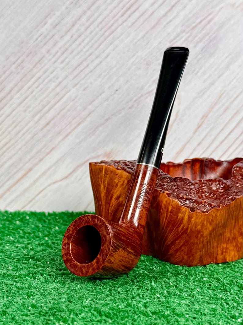 May include: A brown wooden tobacco pipe with a smooth finish and a black stem, featuring the text Kaywoodie stamped on the side. The pipe is positioned in front of a textured, natural-edged wooden bowl with a deep reddish-brown grain.