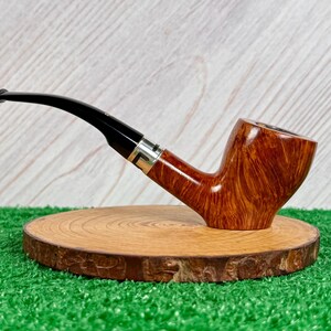 May include: A tobacco pipe with a polished brown wood grain bowl and a curved black stem. The stem features a silver-colored metal band where it connects to the bowl. The pipe is positioned horizontally, showcasing the smooth finish and natural wood texture.